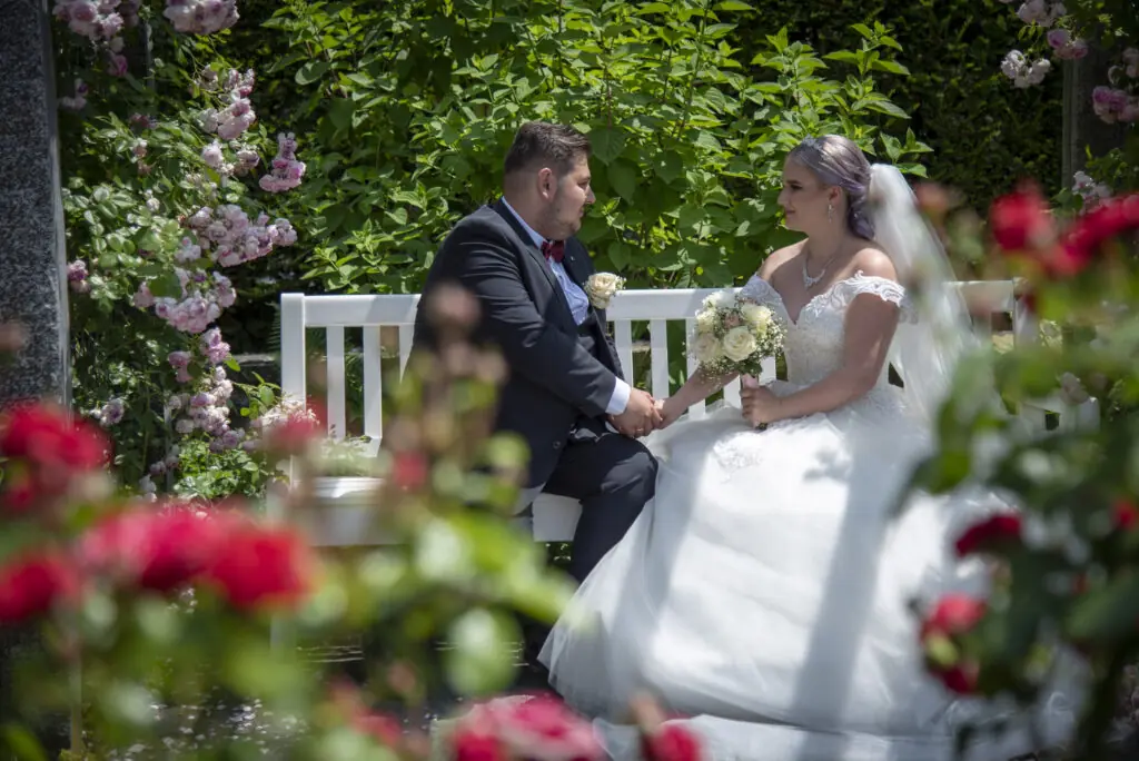 Brautpaar auf weißer Bank im blühenden Rosengarten– emotionale Hochzeitsfotos in Bayern