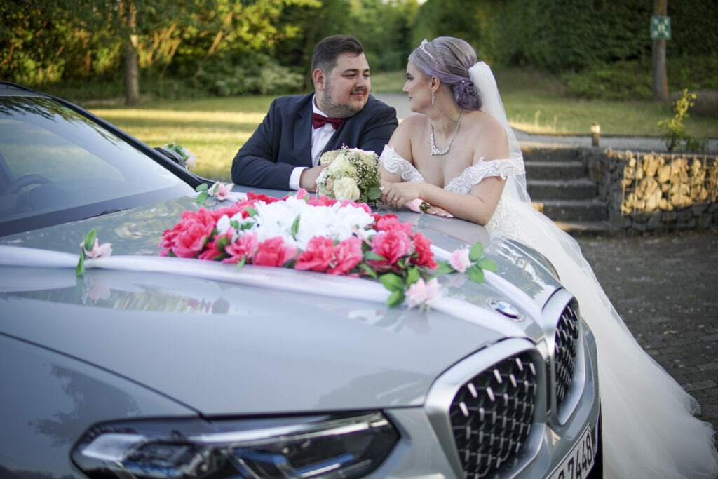 Braut und Bräutigam schauen sich verliebt an hinter geschmücktem Hochzeitsauto – Hochzeitsfotos Alexander Dechant