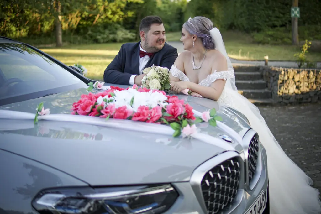 Braut und Bräutigam schauen sich verliebt an hinter geschmücktem Hochzeitsauto – Hochzeitsfotos Alexander Dechant