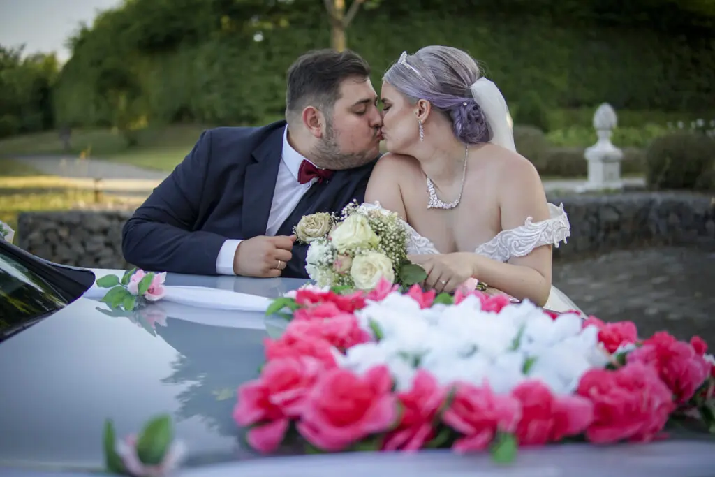 Braut und Bräutigam küssen sich hinter geschmücktem Hochzeitsauto – Hochzeitsfotografie in Bayern
