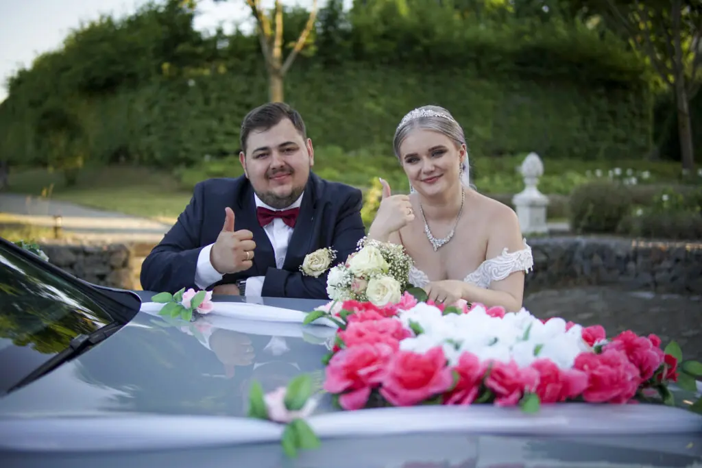 Braut und Bräutigam mit Daumen hoch hinter geschmücktem Hochzeitsauto – Hochzeitsfotograf in Bayern
